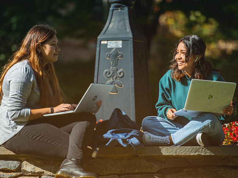 Two students sitting around the campus clock on their laptops